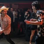 Man wearing a white disco cowboy hat dancing at a lively festival or party with people in sparkly outfits and stylish clothing.